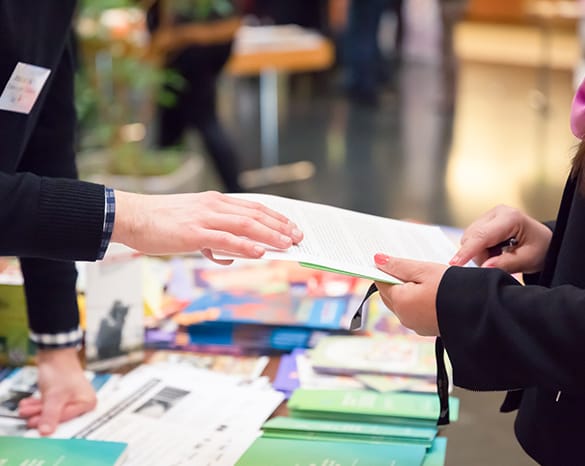 Two people exchanging brochures at a table filled with promotional materials.
