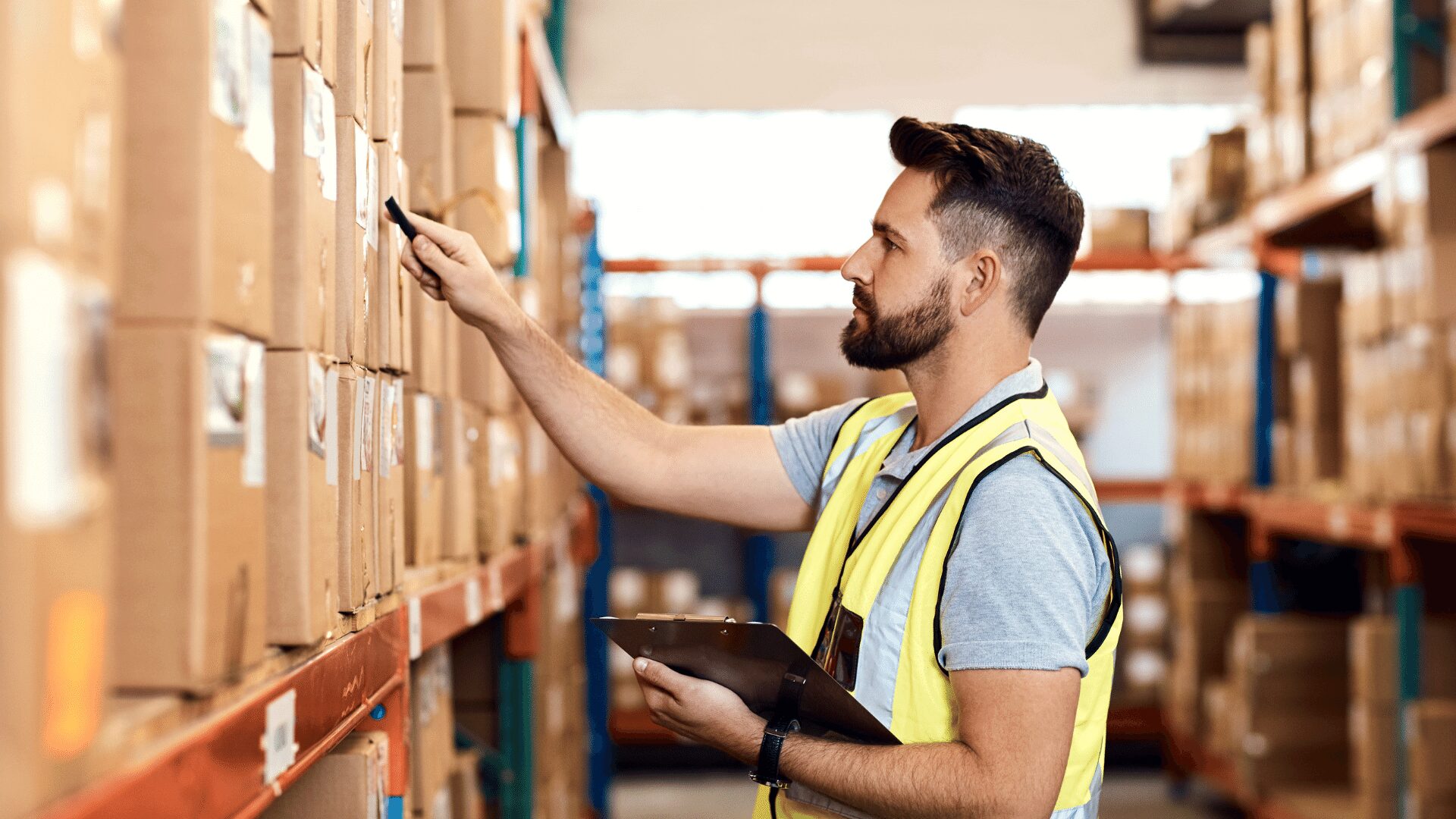 Worker in safety vest scanning boxes in warehouse with clipboard.